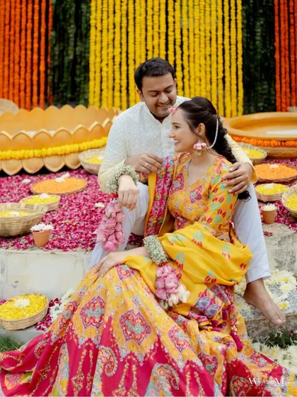 A beautiful portrait of the couple amidst a vibrant Haldi setup. The groom has his arm around the bride, and they are adorned with floral jewelry, creating a picture of love and tradition.