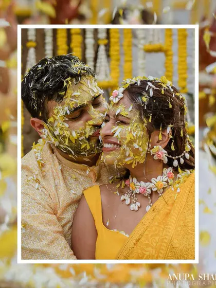 A moment of pure, messy joy during the Haldi. The groom kisses the bride's cheek as they are showered with flower petals, their faces covered in turmeric.