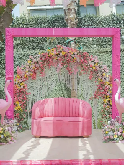 The main seating for the couple at their pastel Haldi. A pink scalloped sofa is set against a backdrop of a floral arch and flanked by two flamingo props, creating a cute and stylish photo spot.