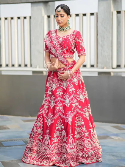 A full-length shot of a bride in a red lehenga with intricate silver floral embroidery, paired with a green choker for a pop of color.