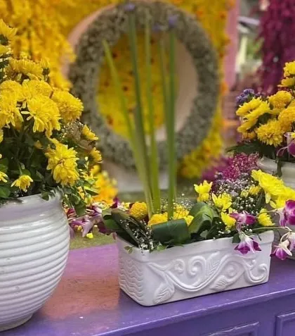 A close-up of a floral centerpiece for a Mehendi or Haldi event. The arrangement includes yellow chrysanthemums and purple orchids in simple white vases.