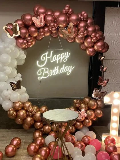 An elegant first birthday setup with a rose gold and white color scheme. The circular balloon arch is accented with butterfly details and surrounds a 'Happy Birthday' neon sign, with a marquee number '1' on the side.