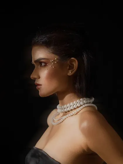 A dramatic, low-key beauty portrait in profile. The lighting highlights the contours of the model's face and the shimmer of the pearl necklace against the dark background.