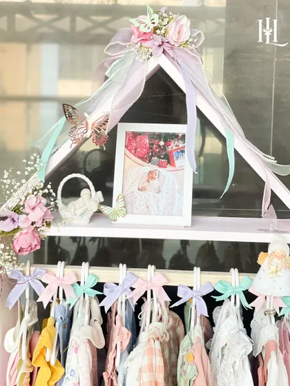 A close-up of the 'Home of Love' baby hamper. This dollhouse-style wardrobe is filled with tiny, adorable baby clothes, a photo frame, and delicate floral and butterfly accents.