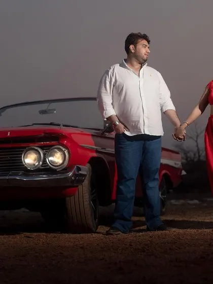 A beautiful evening shot of the couple holding hands in front of a vintage Chevrolet, with the car's headlights on.