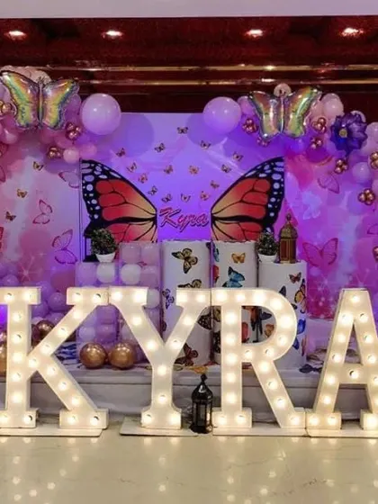 A grand butterfly-themed stage decoration with large marquee letters. The setup includes a purple-lit backdrop and extensive balloon arrangements.