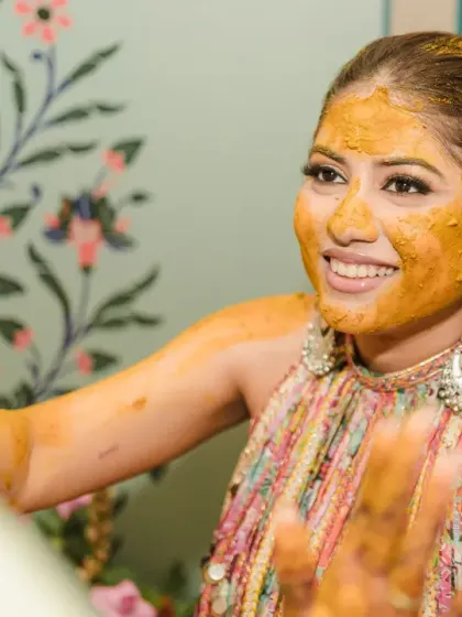 The bride smiles as she participates in the haldi rituals. The soft, painted background and her happy expression create a lovely and elegant portrait.