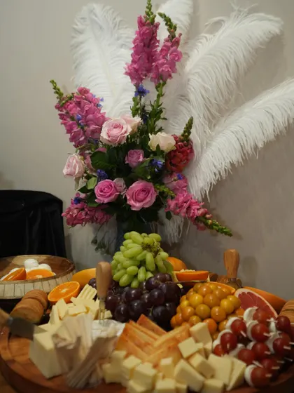 A luxurious cheese board styled with dramatic white feathers and a vibrant floral bouquet. The platter itself is a masterpiece of color and texture, with various cheeses, grapes, and cherry tomatoes.