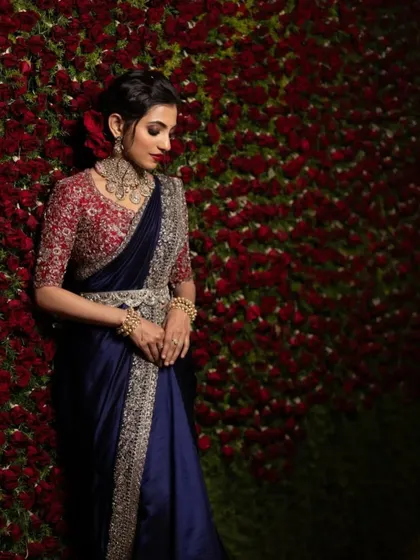 A dramatic and romantic reception look. The bride stands against a wall of red roses, her navy blue saree and bold makeup creating a stunning contrast.