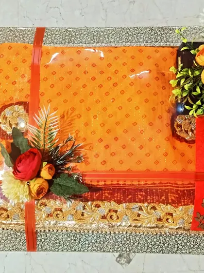 A bright orange and red saree packed in a silver glitter tray, decorated with matching flowers.