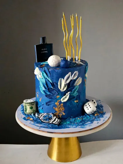 An abstract and stylish cake for him, featuring a textured blue buttercream finish with gold leaf accents. The cake is decorated with edible toppers representing his favorite things, like a watch, perfume, and money.