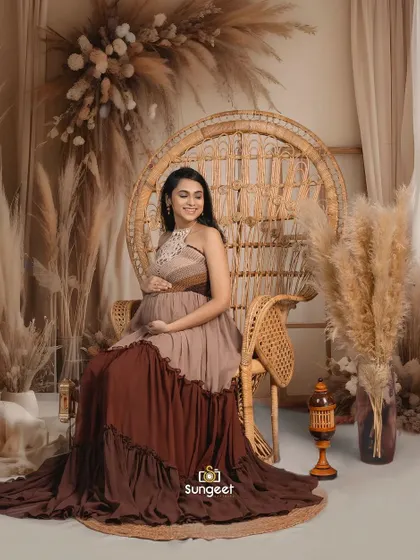 A mom-to-be seated in a large peacock chair, part of our boho-chic collection. The tiered brown dress adds to the earthy, natural vibe.