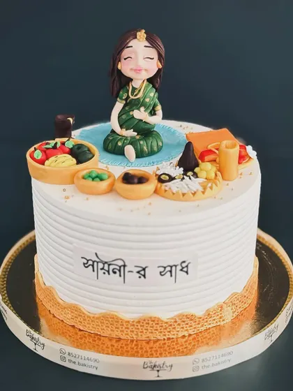 A beautiful and traditional cake for a Bengali baby shower, or 'Shaadh,' featuring a pregnant mother-to-be surrounded by customary foods.