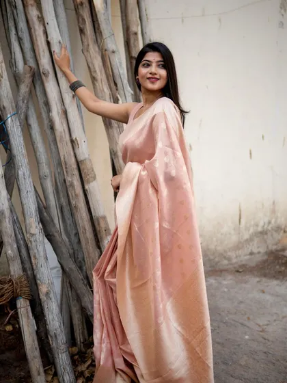 This peach-colored silk saree has a soft, elegant sheen. Posed against a rustic background of wooden logs, the image highlights the saree's delicate color and texture.