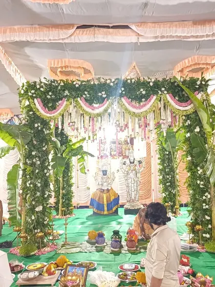 A priest performs rituals at a traditional mandap, with idols of deities and lush green floral decor.