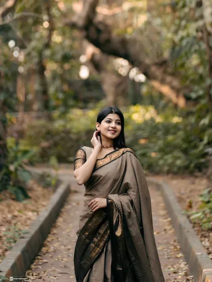 A graceful pose in a muted brown cotton saree with a black and gold border. The natural park pathway and soft lighting create a serene and contemplative mood.