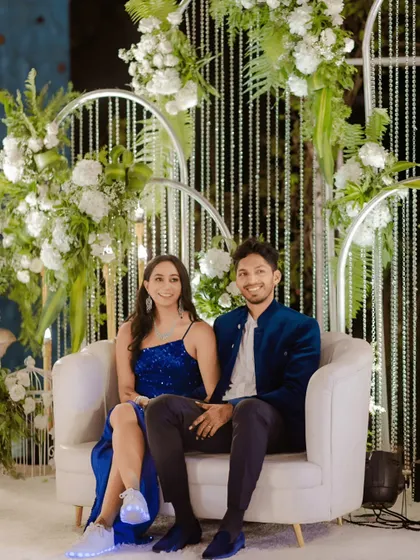 A modern and chic seating area for a Sangeet or reception. The couple is seated on a contemporary white loveseat against a backdrop of cascading crystal strings and fresh white florals, creating an elegant and stylish photo opportunity.