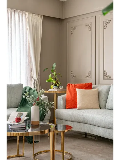 A close-up of the seating area, highlighting the mix of textures and colors. The vibrant orange and soft beige cushions add warmth to the cool grey sofa, complemented by the elegant gold-legged coffee table.
