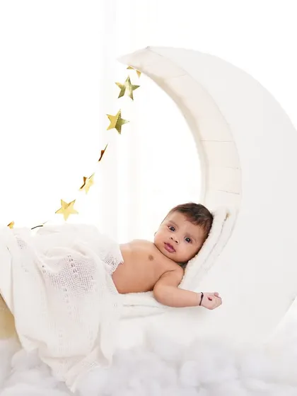A peaceful baby lying on a moon prop under a soft blanket, with a string of golden stars overhead. A magical setup for a baby photoshoot.