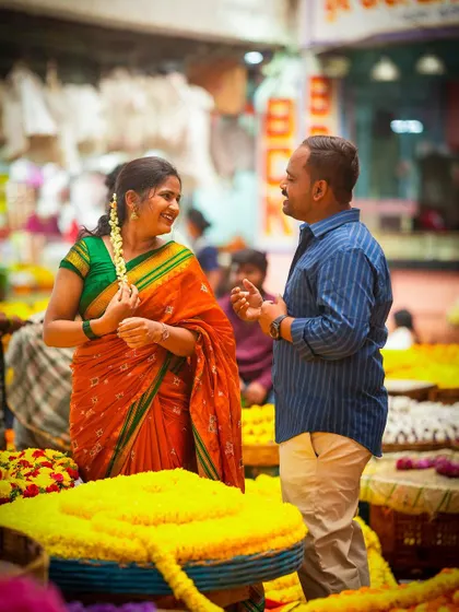 A sweet, interactive moment during a pre-wedding shoot in a flower market, with the groom placing flowers in the bride's hair.