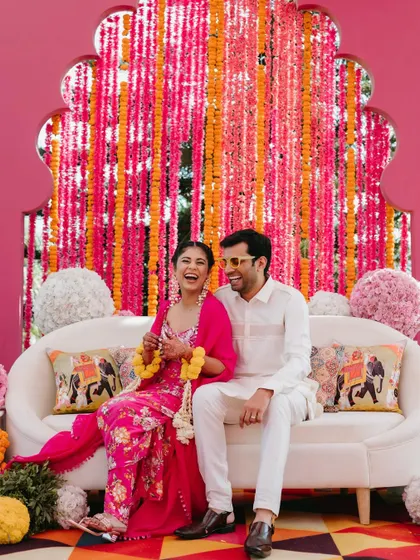 A happy, candid shot of a couple sharing a laugh on a sofa during their Mehendi ceremony, against a vibrant pink and orange floral backdrop.