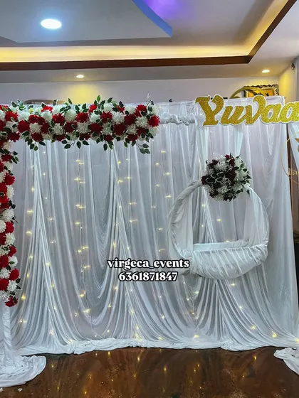 A simple and elegant Naming Ceremony backdrop with white drapes, fairy lights, and a red and white floral arch.