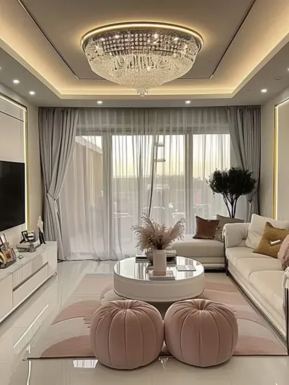 A chic living room with touches of soft pink in the poufs and cushions, adding a gentle warmth to the white and gold theme. A large, ornate chandelier serves as a glamorous centerpiece, elevating the entire space.