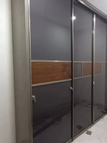 A three-door Aristo sliding wardrobe in dark grey glass with a central wood-finish band. This sleek and modern design offers ample storage while enhancing the room's aesthetic.