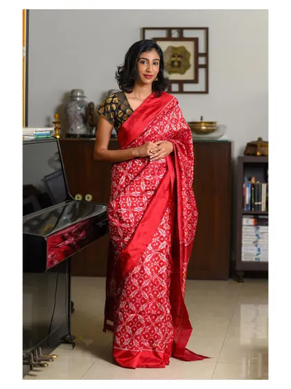 An elegant indoor portrait in a red patola saree. The setting, with the piano and bookshelf, adds a touch of sophistication and context, making it a beautiful lifestyle shot.