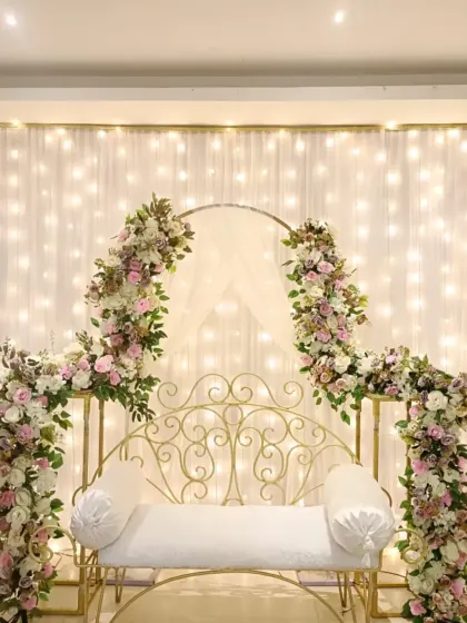 This is a classic and elegant seating arrangement for an engagement or roka. The white wrought-iron bench is framed by beautiful pink and white floral arrangements and a backdrop of warm fairy lights.