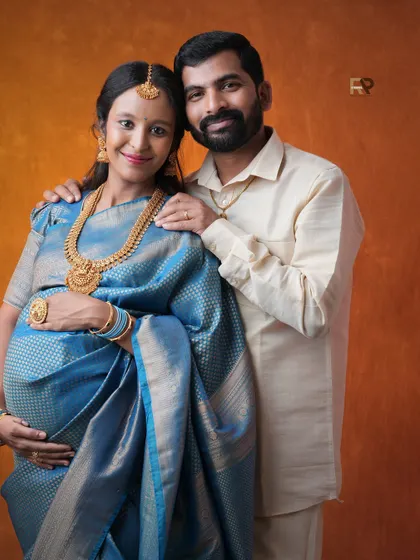 A classic couple's portrait. The parents-to-be look directly at the camera, capturing their shared joy. The rich blue saree and traditional gold jewelry create a striking image.