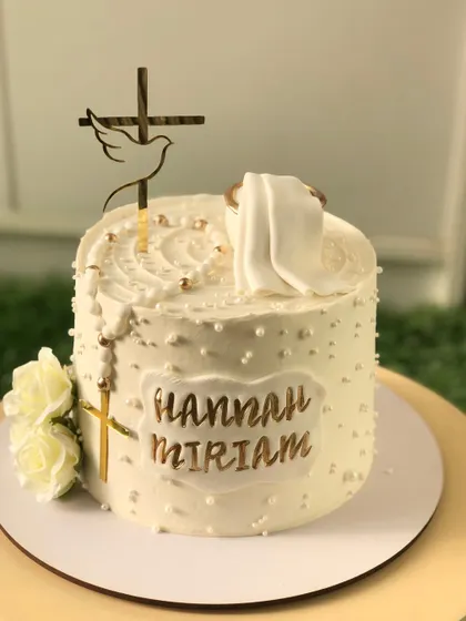 An elegant baptism cake in all white, with a gold cross, a rosary, and the baby's name in gold.