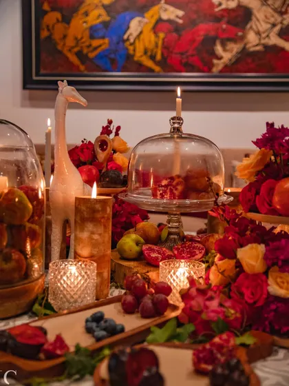 The tablescape is a study in texture and color, with the rich reds of the flowers and pomegranates contrasting with the natural wood and patterned tablecloth. The artwork in the background adds another layer of sophistication.