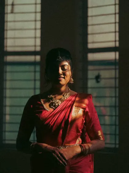 An artistic and moody portrait of a bride in a red saree. The play of shadow and light from the window creates a dramatic, vintage feel, highlighting her silhouette and the richness of her attire.