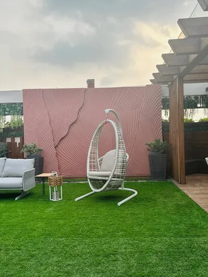 This image highlights the clean lines and modern aesthetic of a terrace project, featuring a comfortable swing chair against a textured pink wall and a well-maintained artificial lawn.