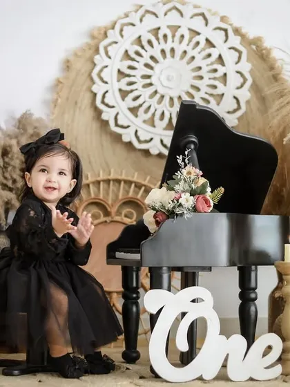 An elegant and stylish first birthday shoot. This little girl, dressed in a beautiful black dress, claps her hands with joy next to a miniature piano in a chic, rustic-themed setup.