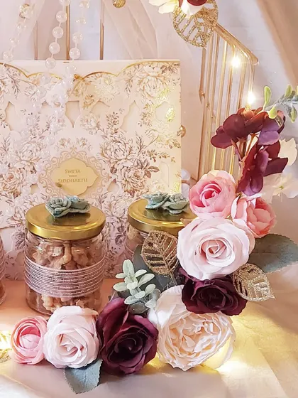 A closer look at the details of our floral wedding invitation hamper. The intricate laser-cut card is the star, complemented by jars of nuts and elegant floral arrangements, all lit by our signature fairy lights.