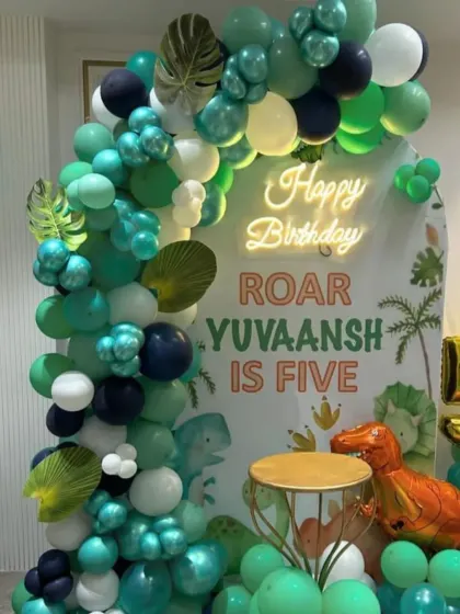 A roaring good time with this dinosaur-themed fifth birthday. The decor includes a custom 'Roar Yuvaansh is Five' backdrop, a green balloon arch with leaf details, and a T-Rex balloon.