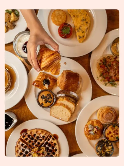 A breakfast feast captured from above, with a hand reaching for a flaky croissant. This lifestyle shot shows a variety of baked goods, from pastries to pancakes, in an inviting, interactive way.
