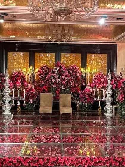 A reception stage drenched in the romance of red. A glass floor filled with rose petals, flanked by grand crystal candelabras and massive floral arrangements, creates an atmosphere of pure opulence.