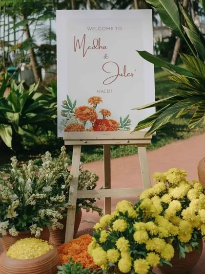 A simple and elegant welcome sign for a Haldi ceremony, surrounded by pots of marigolds and chrysanthemums.