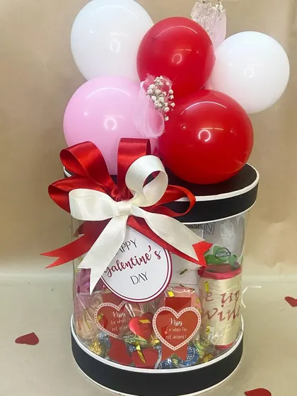 A fun and festive Valentine's hamper in a clear cylindrical box, filled with chocolates and treats and topped with a red, white, and pink balloon bouquet.