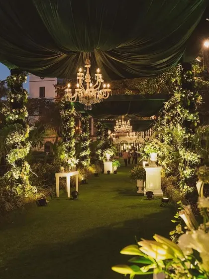 A magical entrance walkway for an evening wedding. The path is lined with lush greenery and white flowers, with elegant chandeliers and green velvet drapes overhead creating a dramatic and enchanting effect.