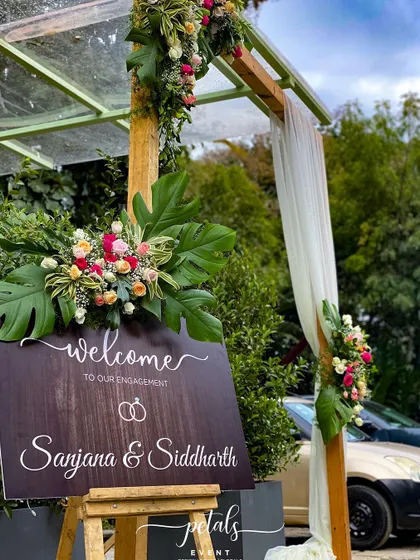 An engagement entrance adorned with pastel roses, tropical leaves, and a wooden sign. The combination of soft colors and natural textures creates a romantic and welcoming atmosphere.