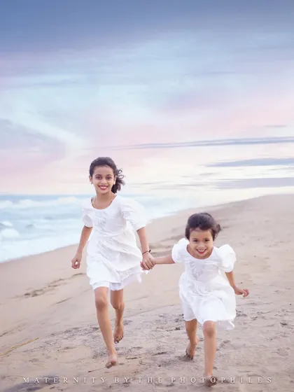 Two sisters holding hands and running freely on the sand. This sibling photograph captures the carefree spirit of childhood against the beautiful backdrop of the sea and a soft, painted sky.
