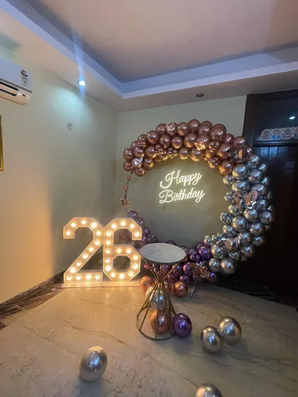 A stylish half-arch balloon ring for a 26th birthday. This home decoration uses chrome rose gold and silver balloons, a neon sign, and marquee numbers.