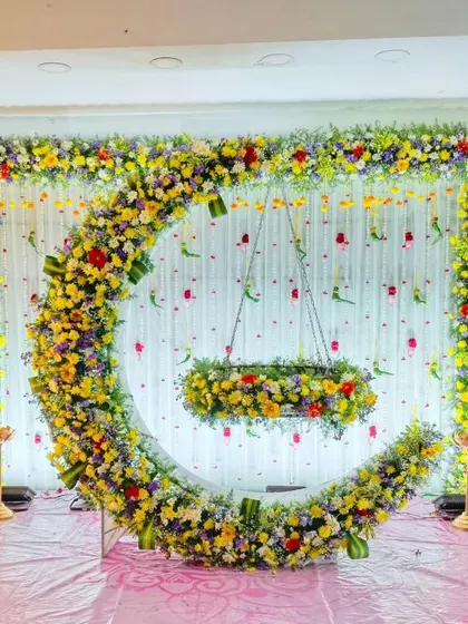 A vibrant and colorful cradle ceremony decoration. The large floral crescent moon is made from a mix of yellow, purple, and red flowers, creating a joyful and celebratory atmosphere.