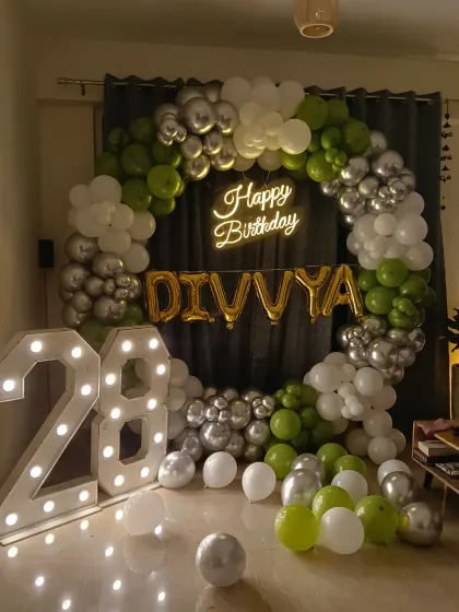 A 28th birthday party with a circular balloon arch in green, silver, and white, complemented by a neon sign and large marquee numbers.