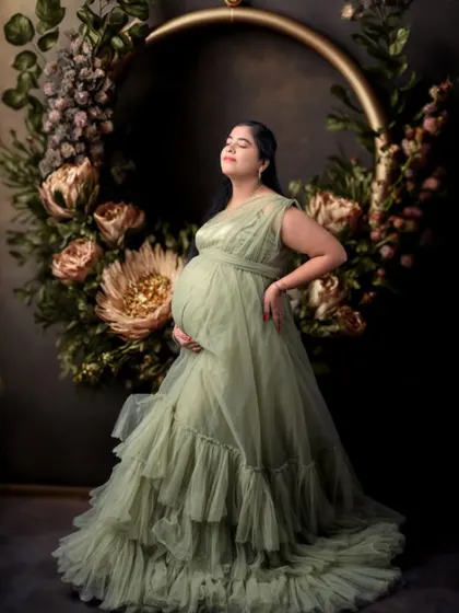 This artistic portrait features a sage green gown and a large floral hoop prop. The dark, moody background makes the subject and the florals stand out.