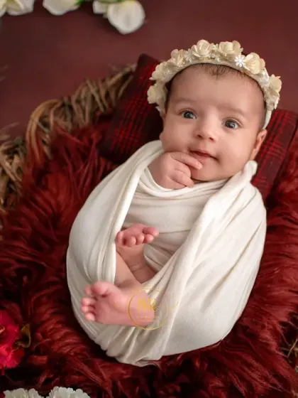 An alert newborn wrapped in white, looking curiously at the camera while nestled in a basket surrounded by red and white flowers.